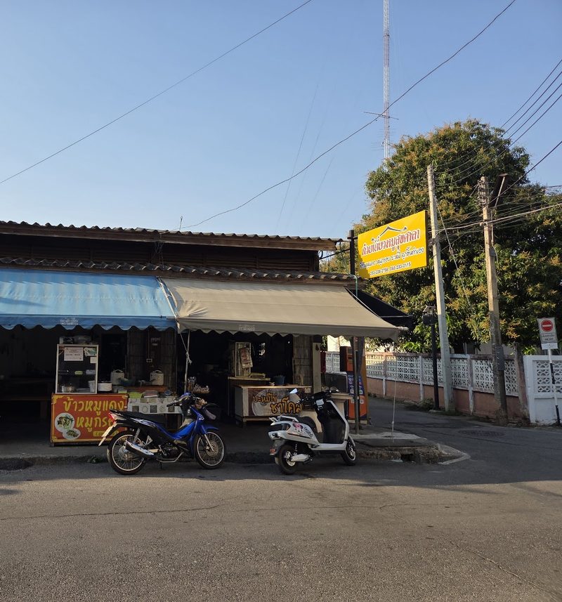 หน้าร้านในซอย ป้ายเหลืองชื่อก๋วยเตี๋ยวหมูสุขศาลา กันสาดผ้าใบ และป้ายรถเข็นข้าวหมูแดงหมูกรอบกับบะหมี่ไข่ชาโคล