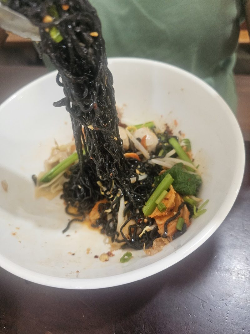 คีบบะหมี่ดำขึ้นจากชาม เห็นผักและพริกอยู่ก้นชาม