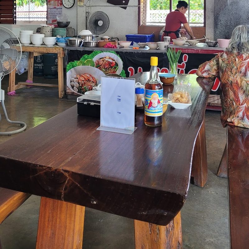 โต๊ะไม้ตัวหนาในร้าน มีขวดเครื่องปรุงวางบนโต๊ะ และเคาน์เตอร์ครัวด้านหลัง