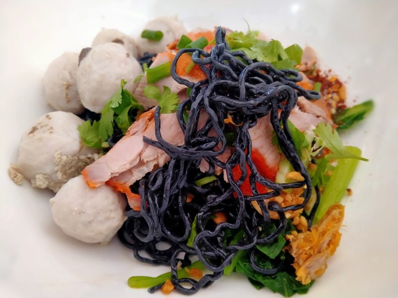 บะหมี่ชาโคลเส้นดำแห้ง ใส่ลูกชิ้นหมู หมูแดง ผัก และพริก ในชามขาว